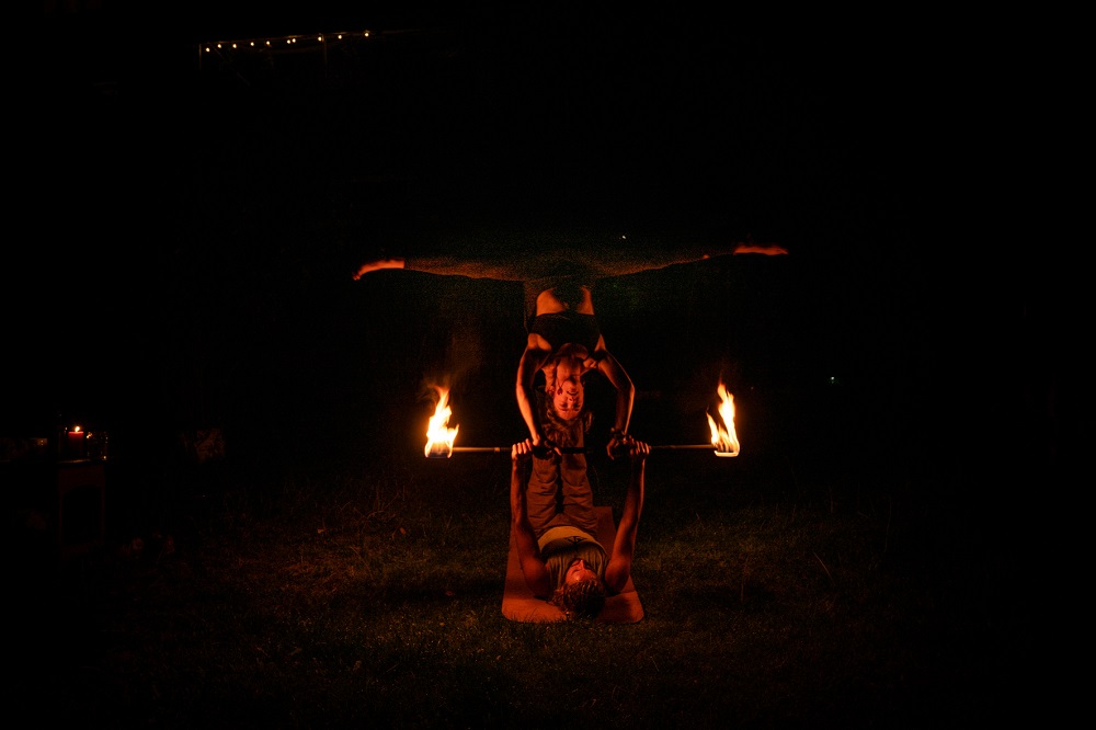 AcroYoga star pose: a flyer balances horizontally on the base's feet with a fire staff held between them