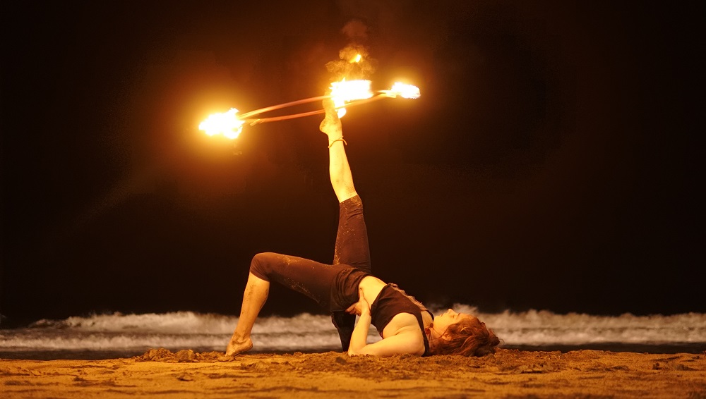 Fire performer balancing a burning fire hula hoop on her foot on a beach at night