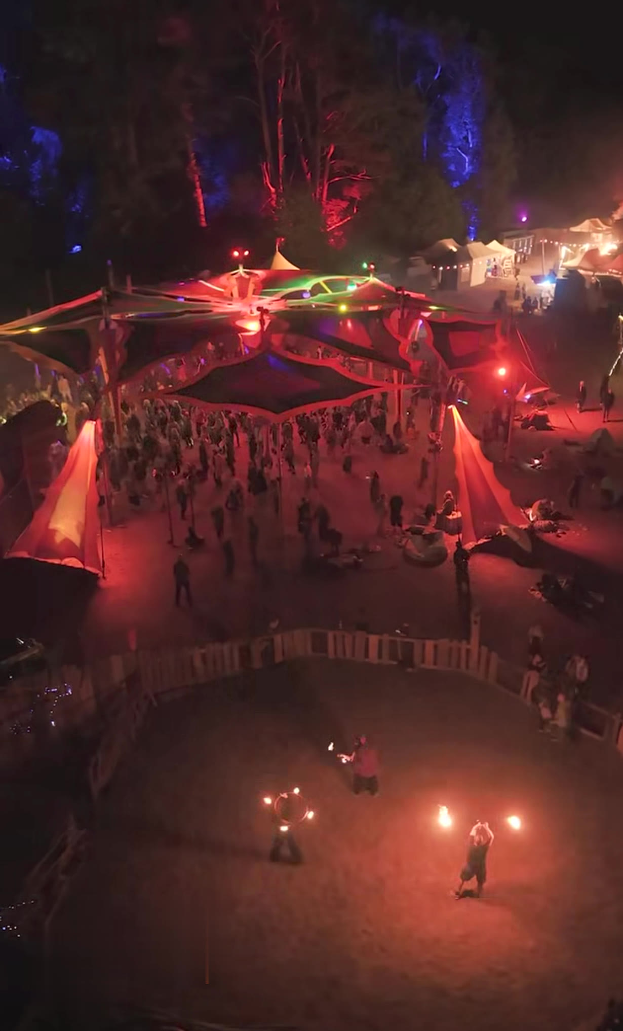 Aerial view of the Firespace at the New Healing Festival at night: fire performers in a fenced fire circle, behind it a large sail-covered dance floor with festival guests